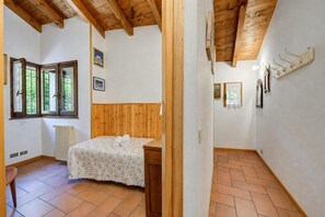 A bedroom with a wooden headboard, a bed with a floral bedspread, a radiator, and a window with a view of trees. - Villa Il Castagno: Few min From Beach, 2BR (Castelveccana)