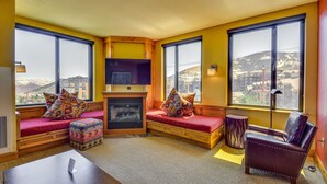 A cozy room with a fireplace, large windows, a sofa, and a TV. - Park City Condo: Steps to Canyons Village & Skiing (Park City)