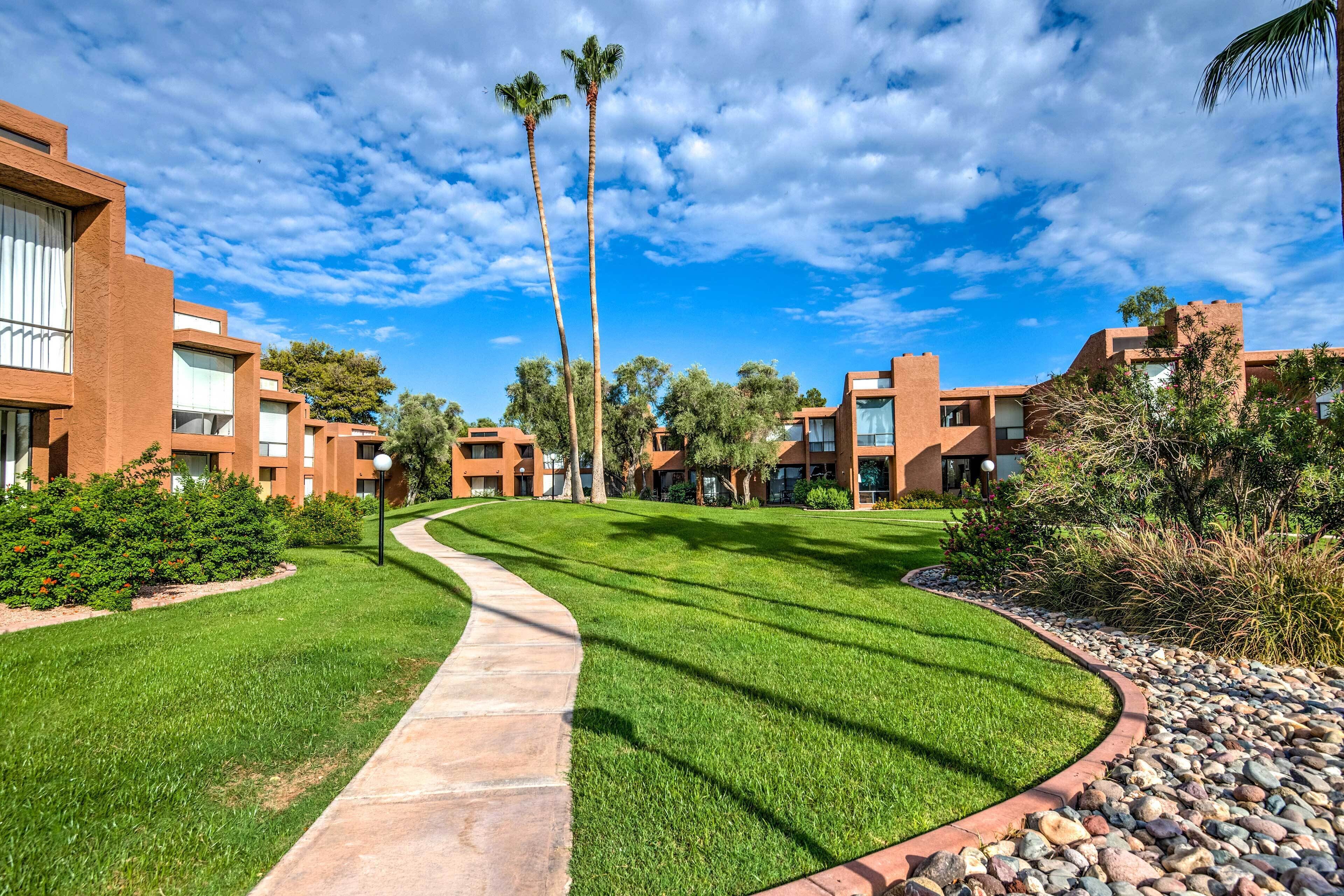 A modern building complex with a central pathway, surrounded by well-maintained lawns and palm trees.