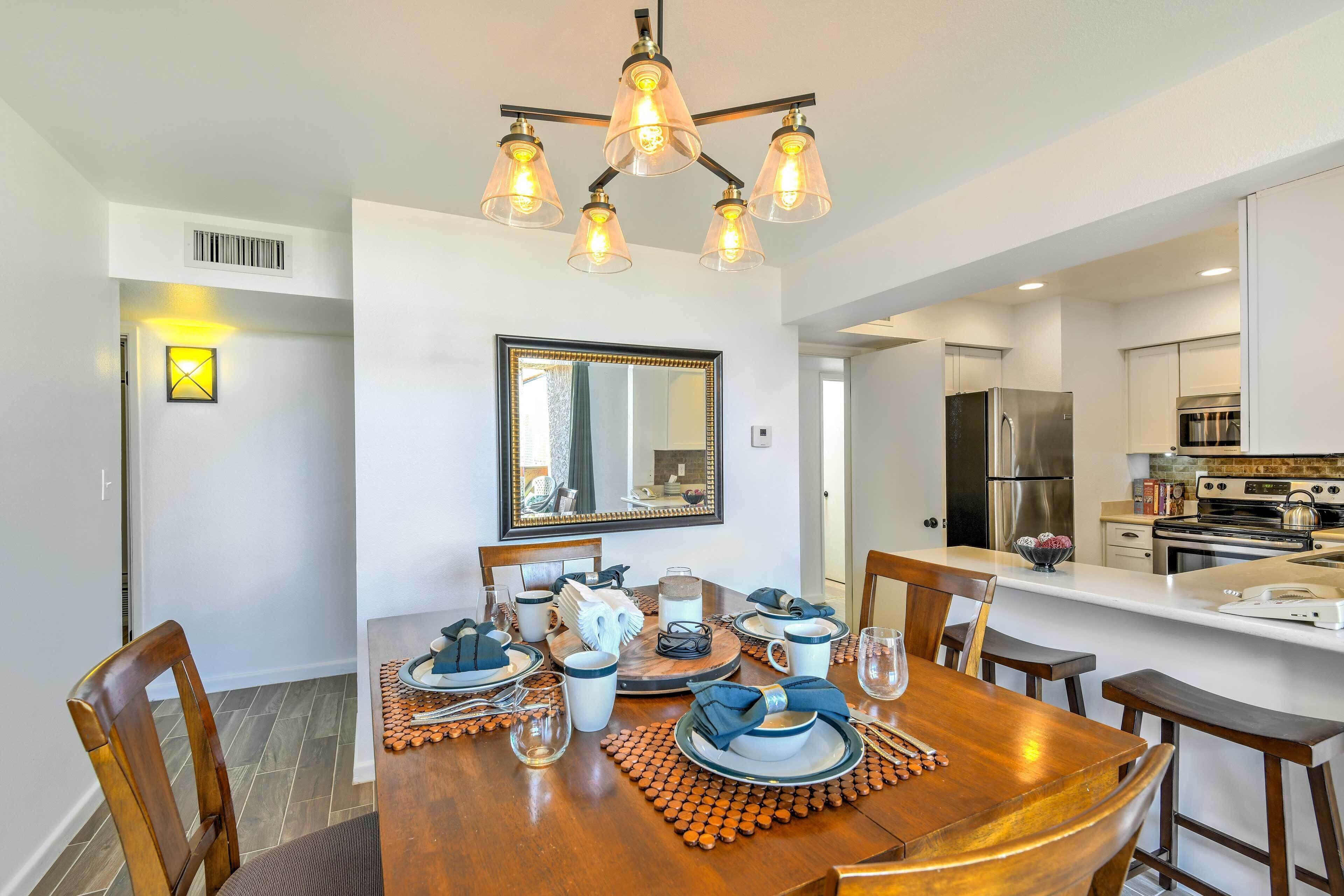 A dining area with a wooden table set for four, a chandelier, and a kitchen in the background.