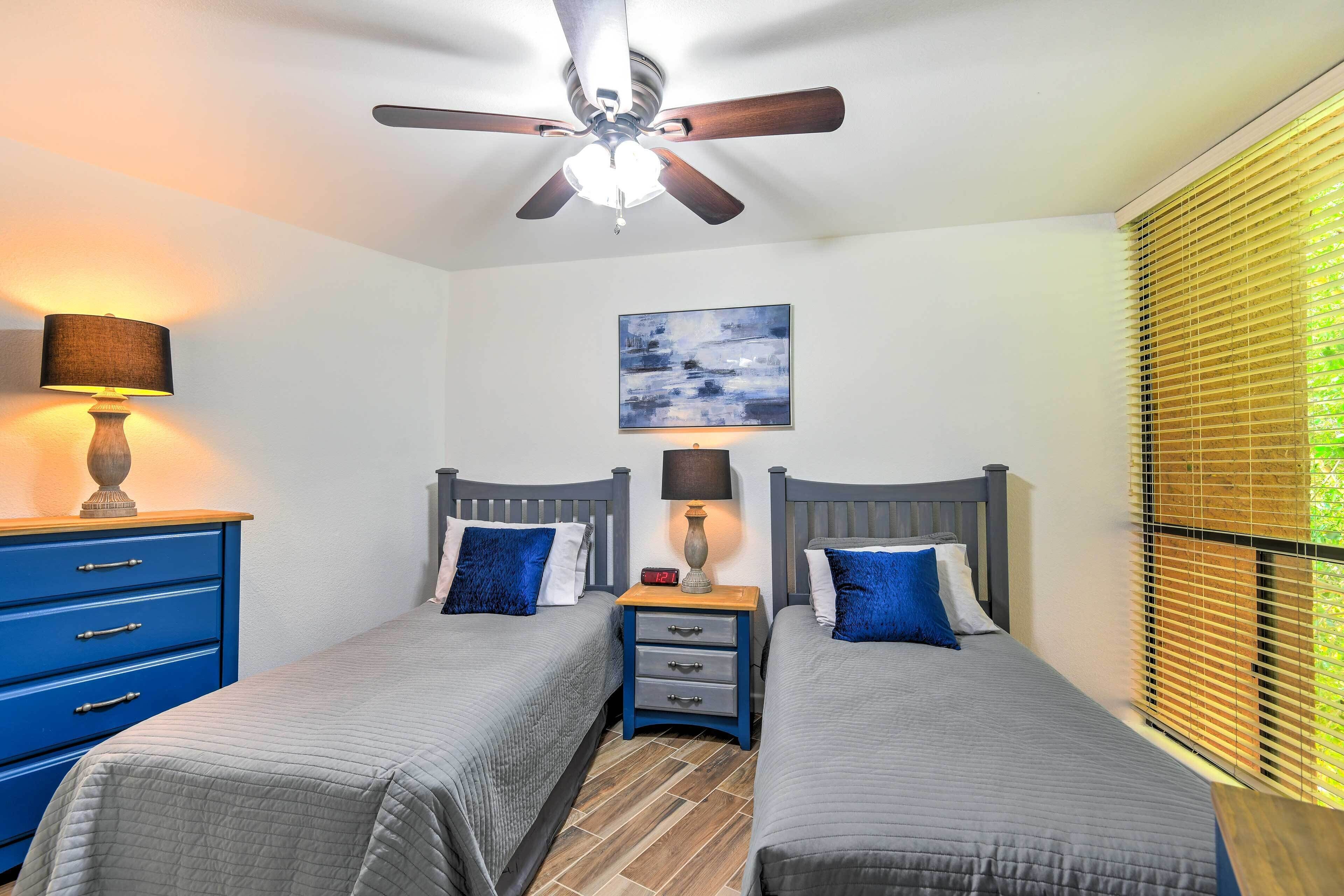 A bedroom with two beds, a ceiling fan, a nightstand with a lamp, a blue dresser, and a window with blinds.