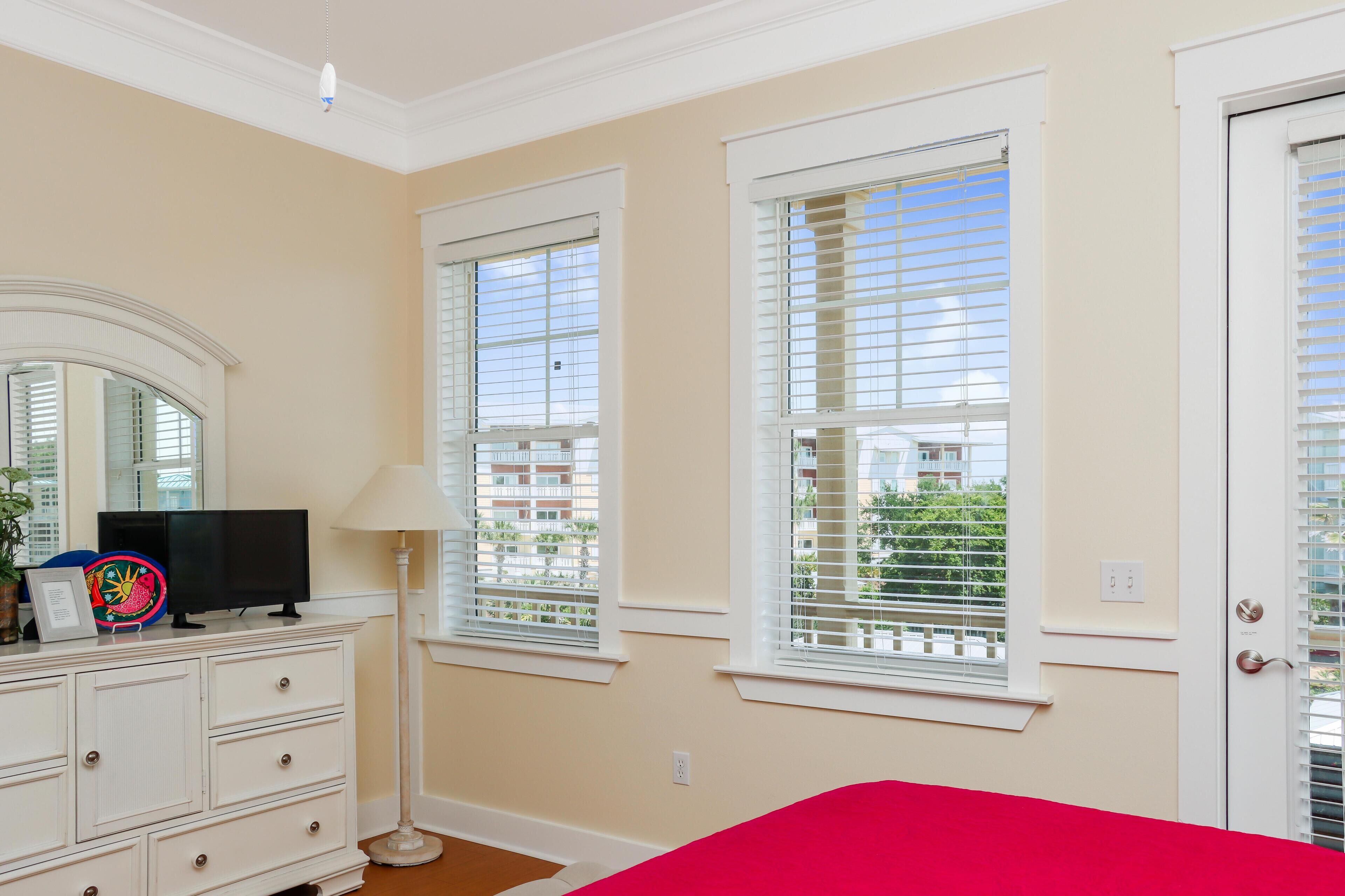 A bedroom with a large window, a dresser, a television, and a lamp.