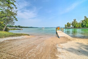 A beach with a wooden pier extending into calm waters, surrounded by greenery and a clear sky.