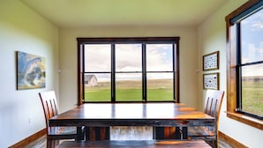 Un comedor con una gran mesa de madera y sillas, vista a un campo de césped a través de amplias ventanas, y obras de arte en las paredes.