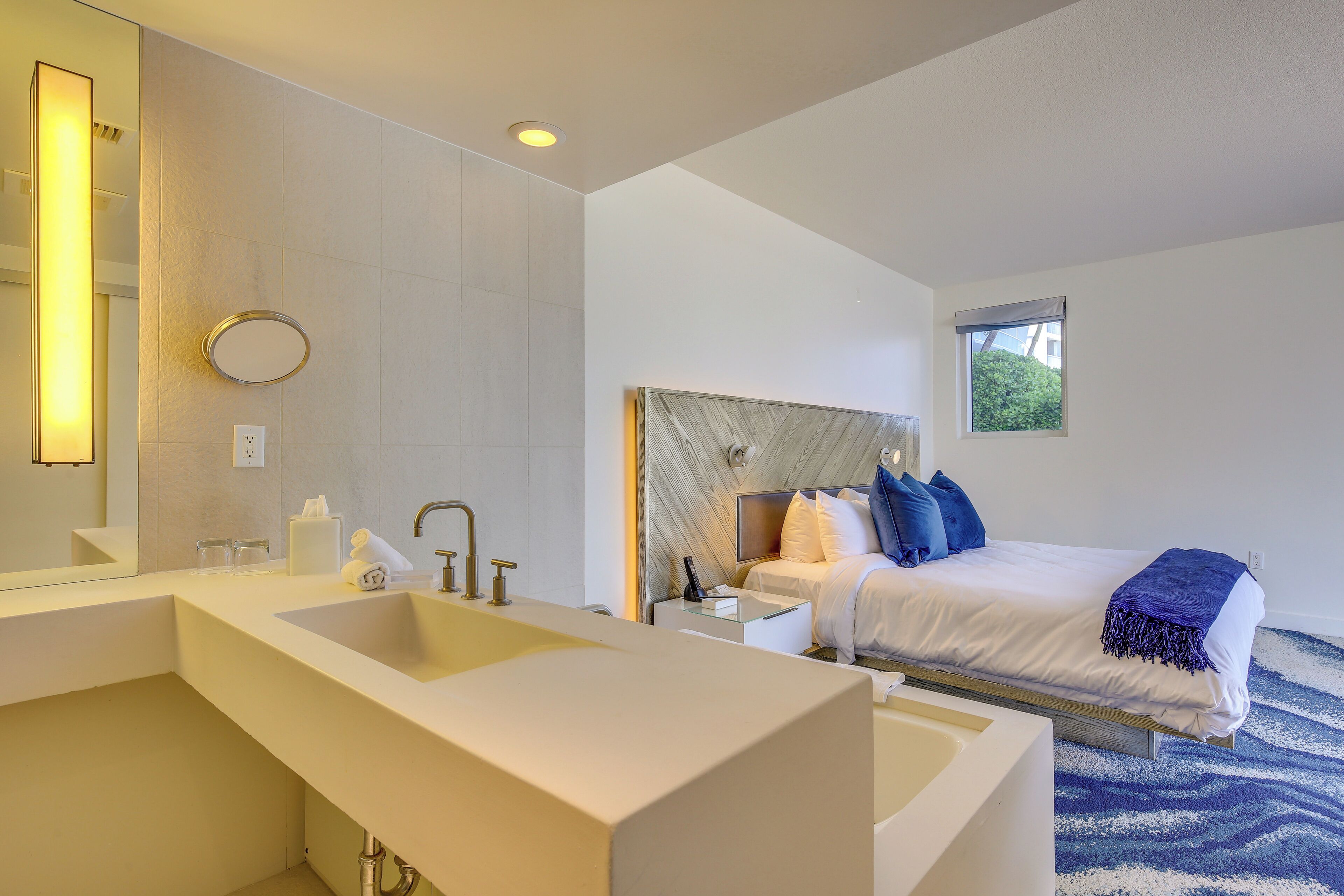 Habitación de hotel moderna con un lavamanos grande en el baño, una cama con cabecero y una ventana con vistas a zonas verdes.