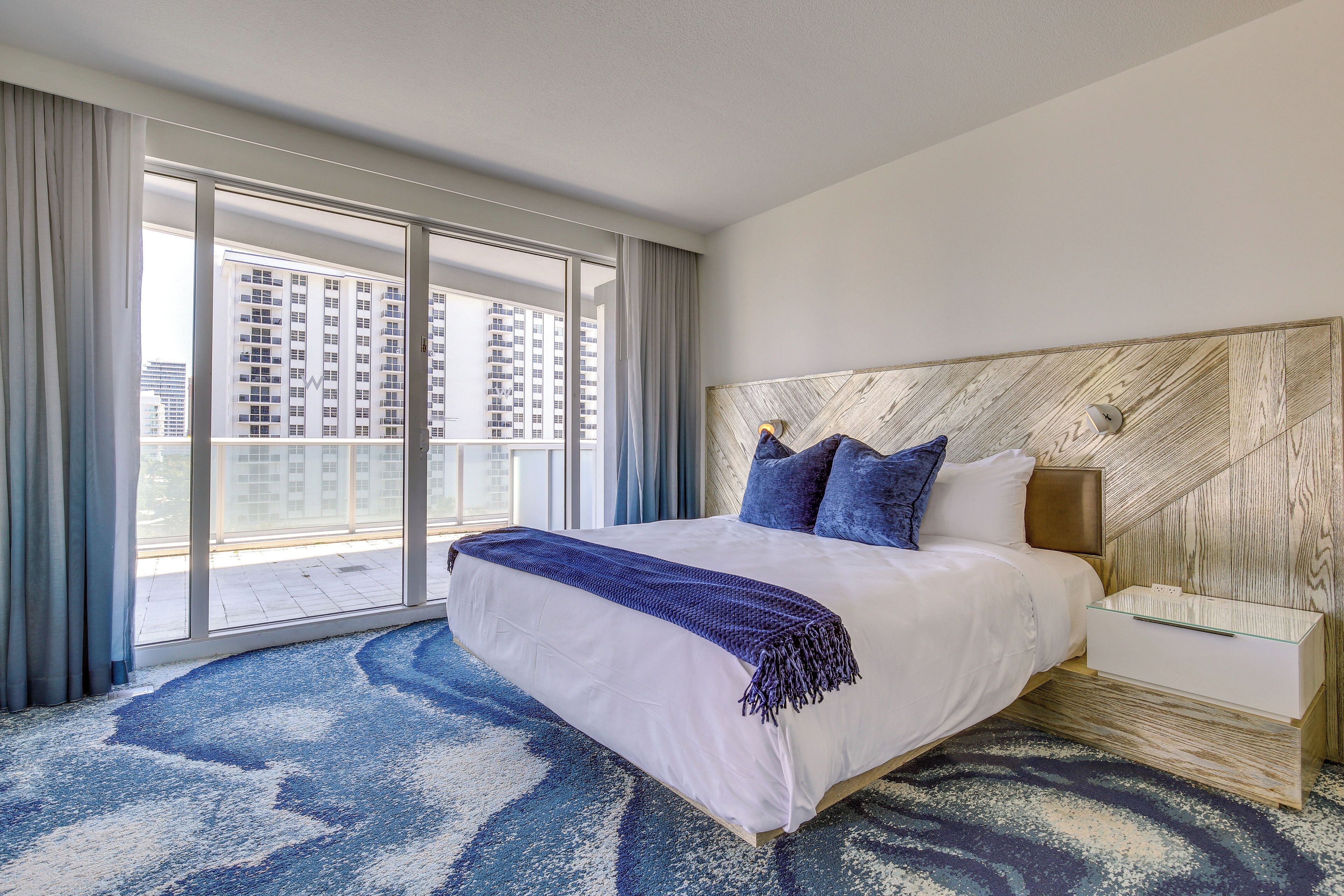 A hotel room with a large bed, a wooden headboard, and a view of tall buildings through floor-to-ceiling windows.