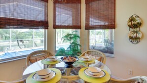 A dining area with a round table set for four, surrounded by wicker chairs. There are three windows with brown blinds, a potted plant in the center, and a decorative wall piece above the table. - Near Sand & Sun, Cape Coral Home w/ Private Pool (Cape Coral)
