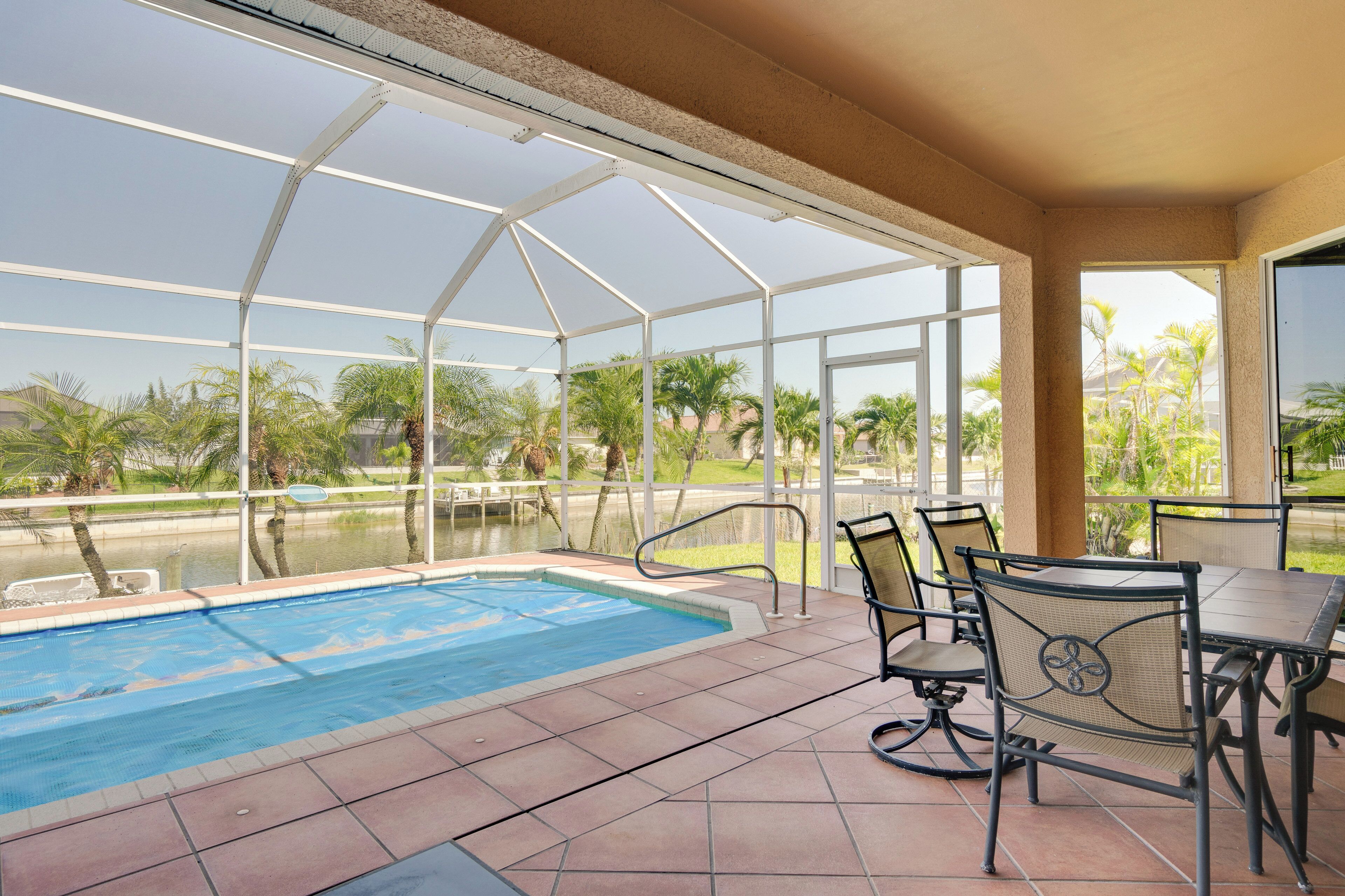 A covered outdoor pool area with a table and chairs, surrounded by glass panels and palm trees.