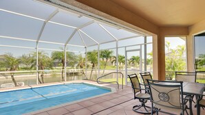 A covered outdoor pool area with a table and chairs, surrounded by glass panels and palm trees. - Near Sand & Sun, Cape Coral Home w/ Private Pool (Cape Coral)
