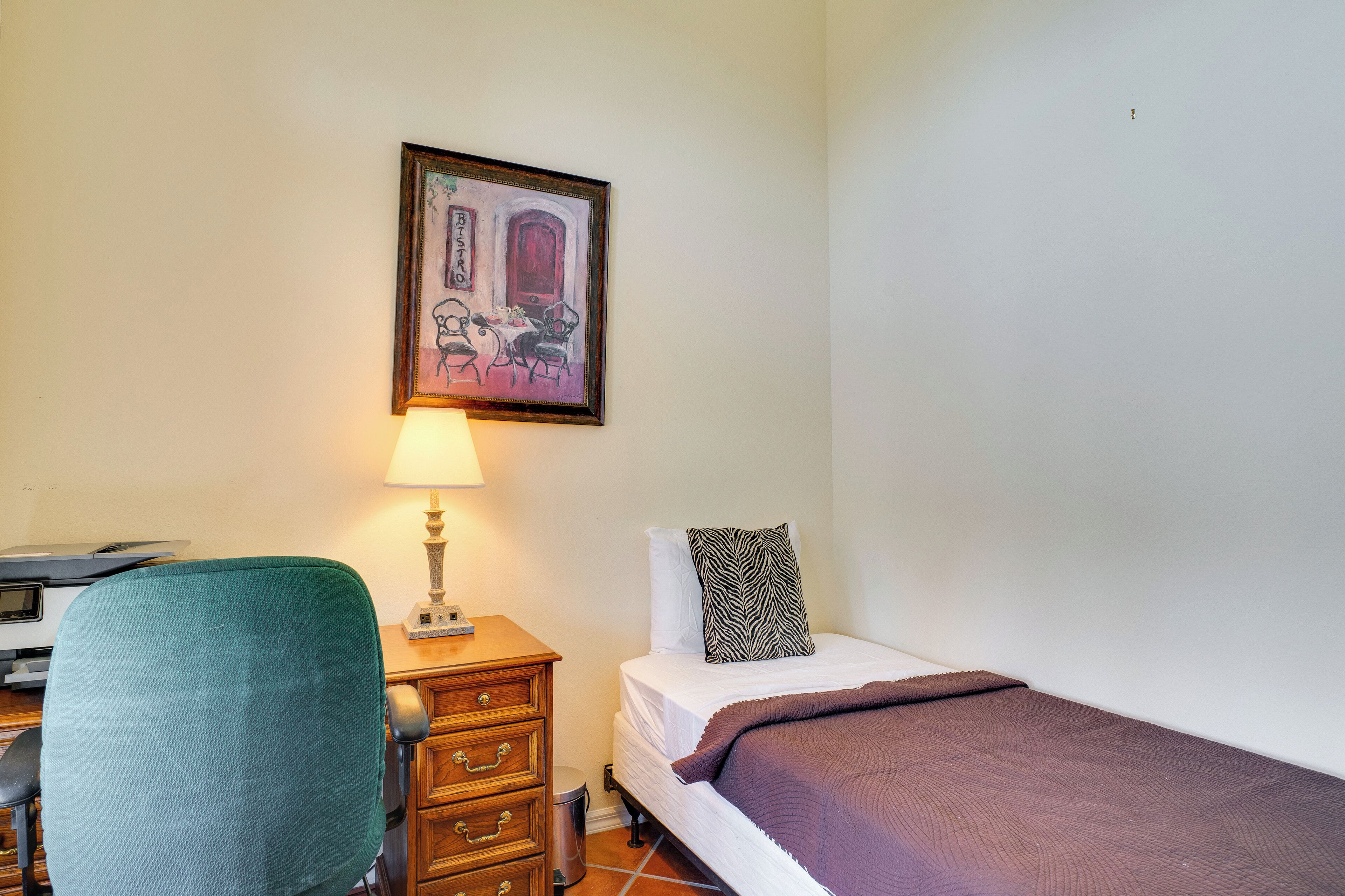 Une chambre à coucher avec un lit, un bureau avec une chaise, une lampe, une poubelle et un tableau encadré au mur.