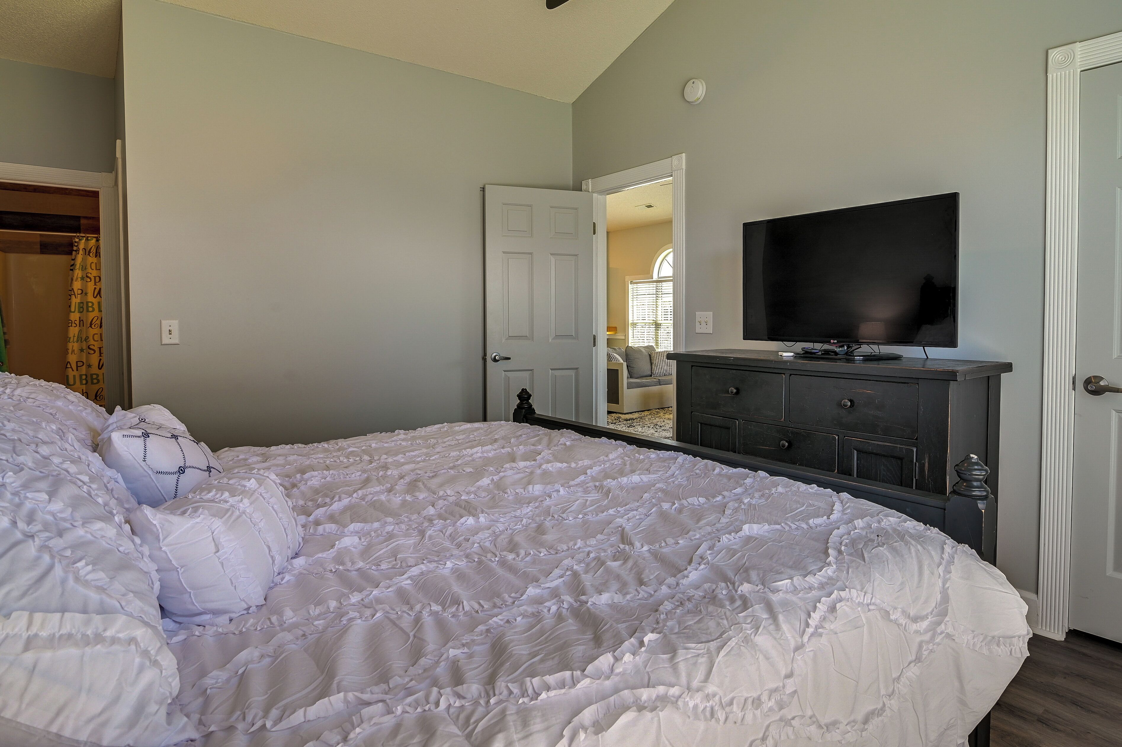 A bedroom with a large bed, a dresser, a television, and a door leading to another room.