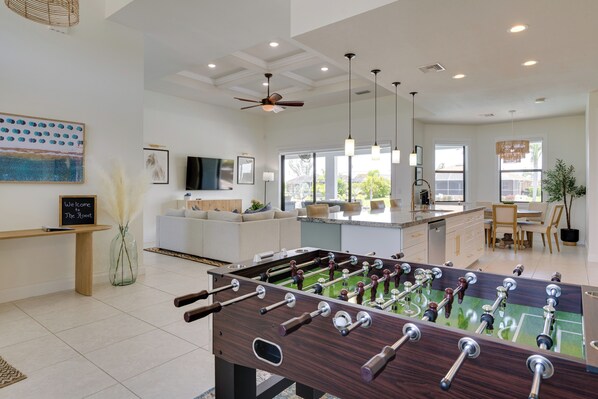 A modern living room with a foosball table, a sofa, a TV, and a dining area with a chandelier.
