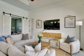 A modern living room with a sectional sofa, a round coffee table, and a white armchair. There is a wooden cabinet below a flat-screen TV, and a floor lamp is visible.