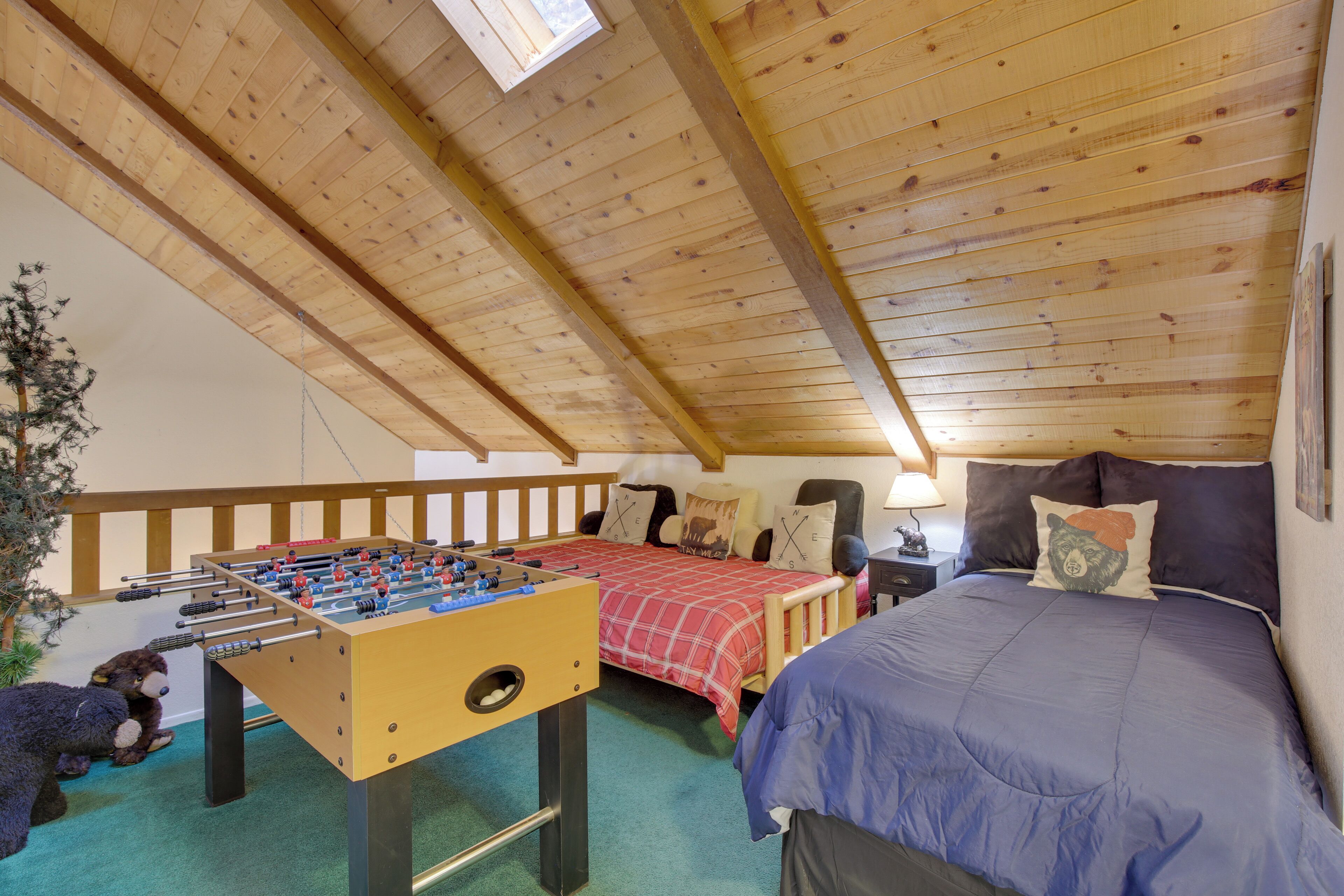 Une pièce chaleureuse avec un plafond en bois, une table de baby-foot, un lit recouvert d’une couverture bleue et un petit bureau avec une lampe.