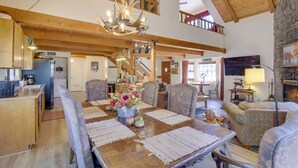 A spacious dining area with a wooden table set for six, surrounded by upholstered chairs. A chandelier hangs from the ceiling, and a staircase leads to an upper level. - Dancing Bear Lodge ~ 1 Mi to Big Bear Lake! (Big Bear Lake)