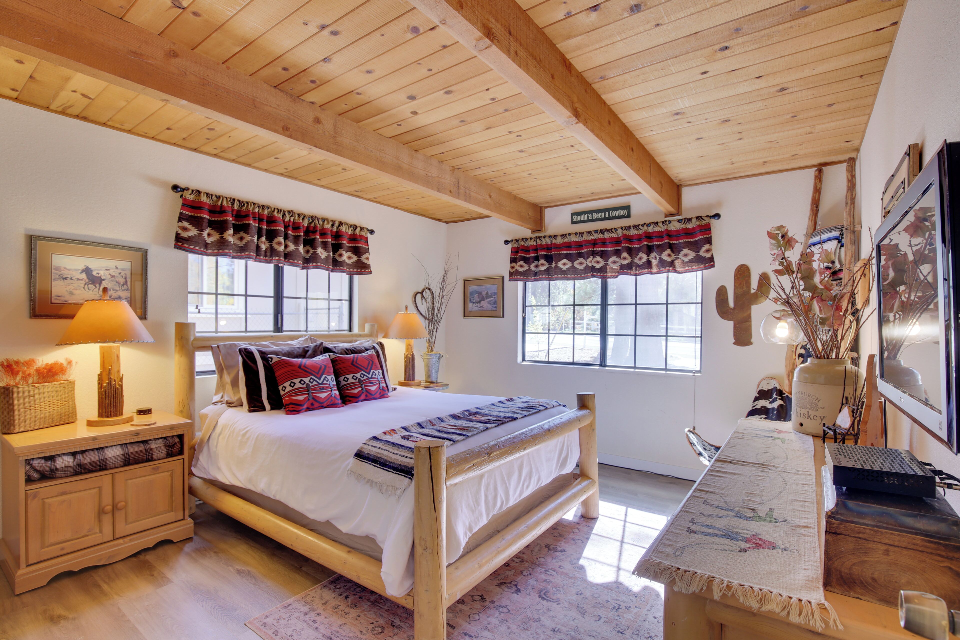 A bedroom with a wooden bed, two bedside tables with lamps, a desk with a computer, and a window with a view of the outside.