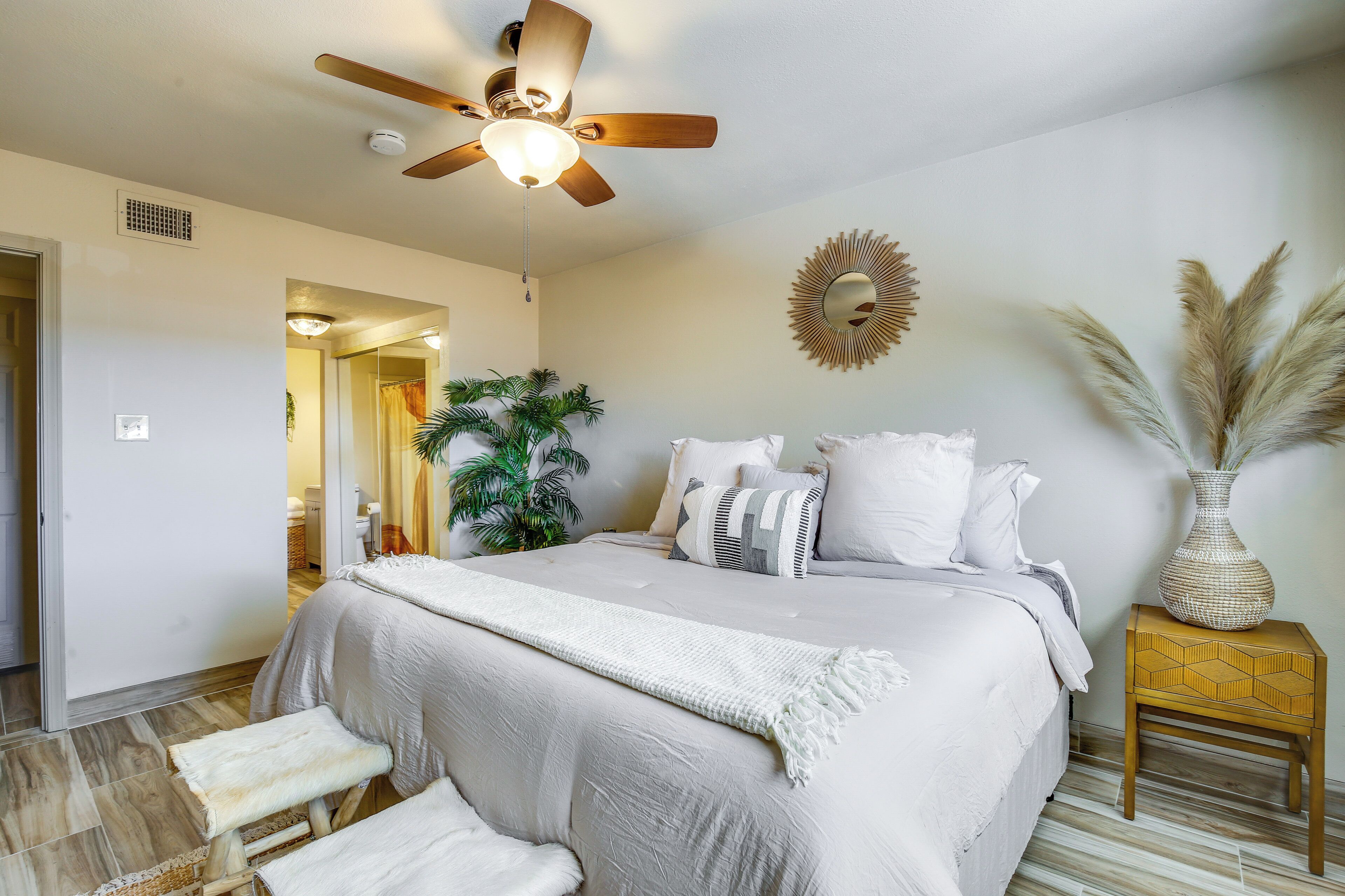 A bedroom with a bed, a ceiling fan, a bedside table with a vase, and a wall-mounted mirror.