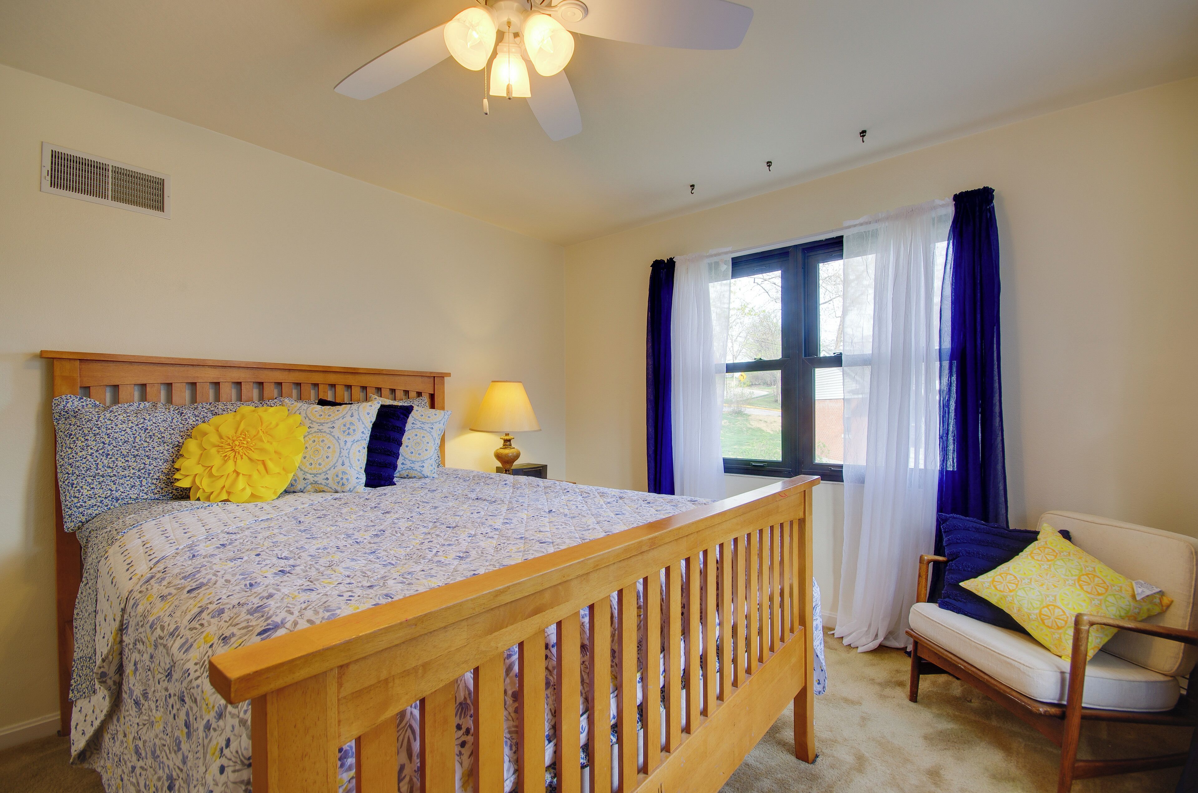 A bedroom with a wooden bed, a nightstand with a lamp, a window with blue curtains, and a chair with a cushion.