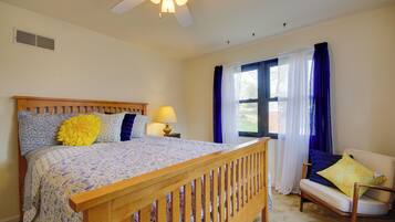 A bedroom with a wooden bed, a nightstand with a lamp, a window with blue curtains, and a chair with a cushion.