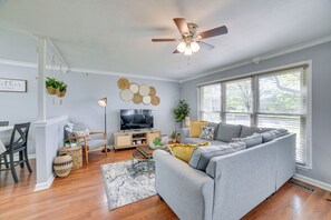 Ein Wohnzimmer mit grauem Sofa, Holzboden, Deckenventilator, Fernseher und einem Essbereich mit Tisch und Stühlen.