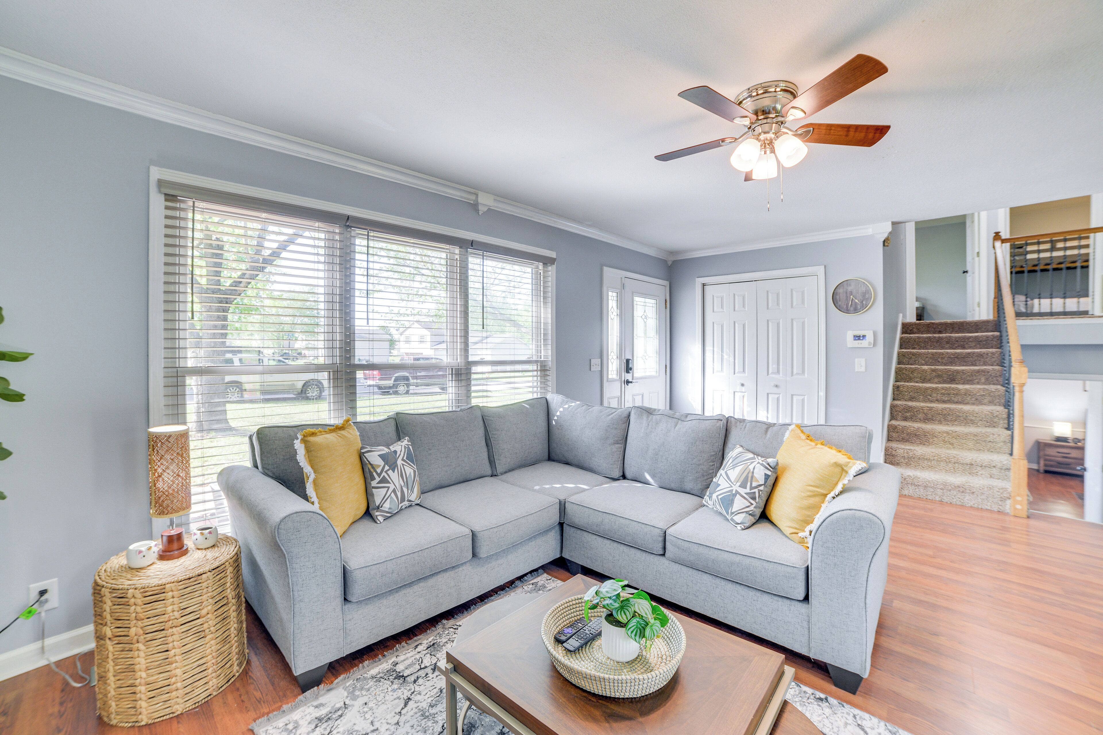 A living room with a gray sectional sofa, a ceiling fan, and a staircase in the background.