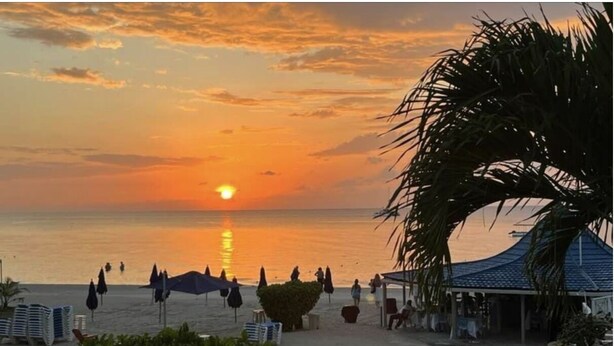 A beach scene with a sunset, umbrellas, and a pathway leading to a building. - Seacret Studios - Cozy Jamaican Retreat Apts by RedAwning (Negril)