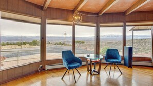 Een kamer met grote ramen, twee blauwe stoelen, een glazen tafel en een moderne kroonluchter.