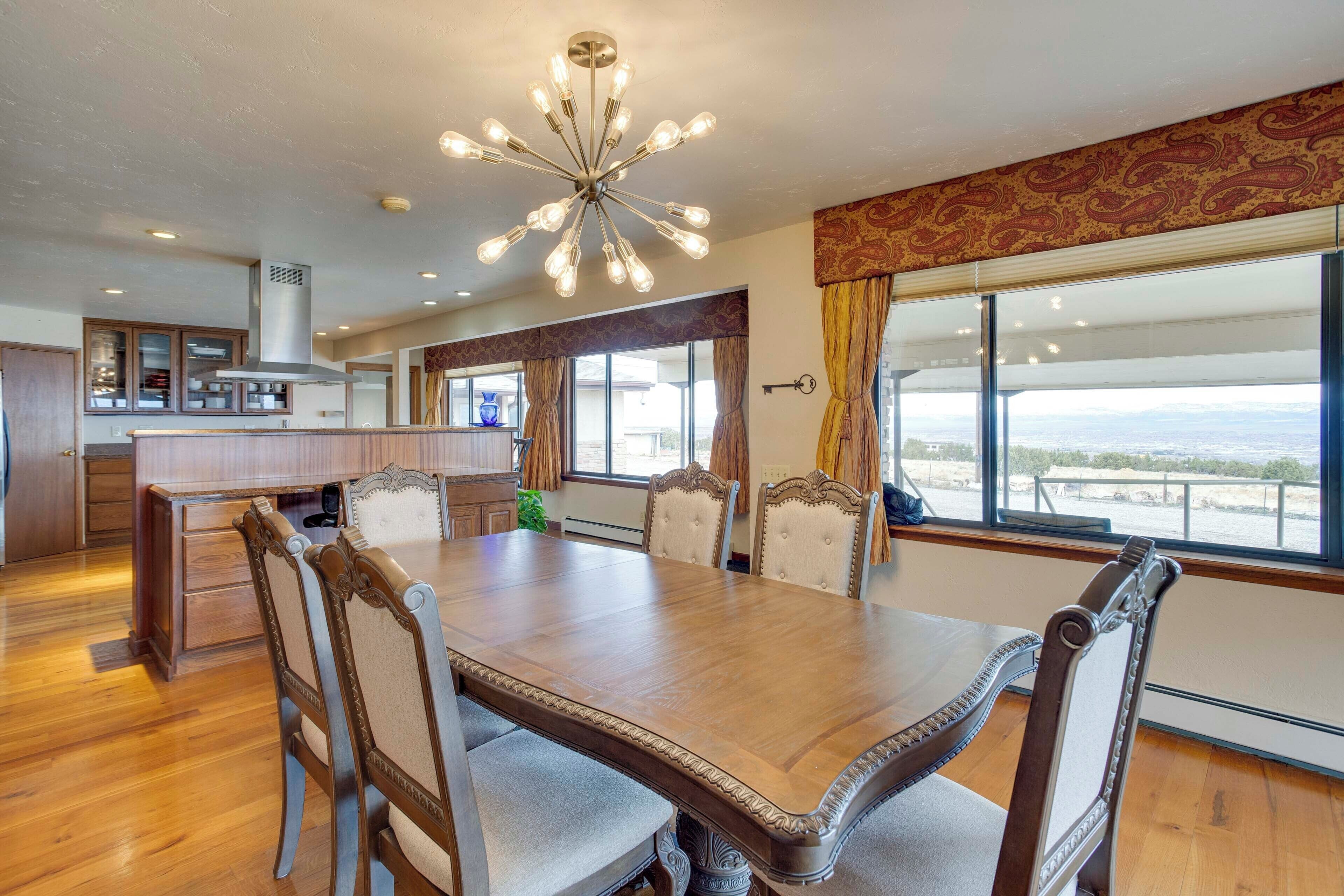 A dining area with a large wooden table and chairs, a chandelier, and large windows with a view.