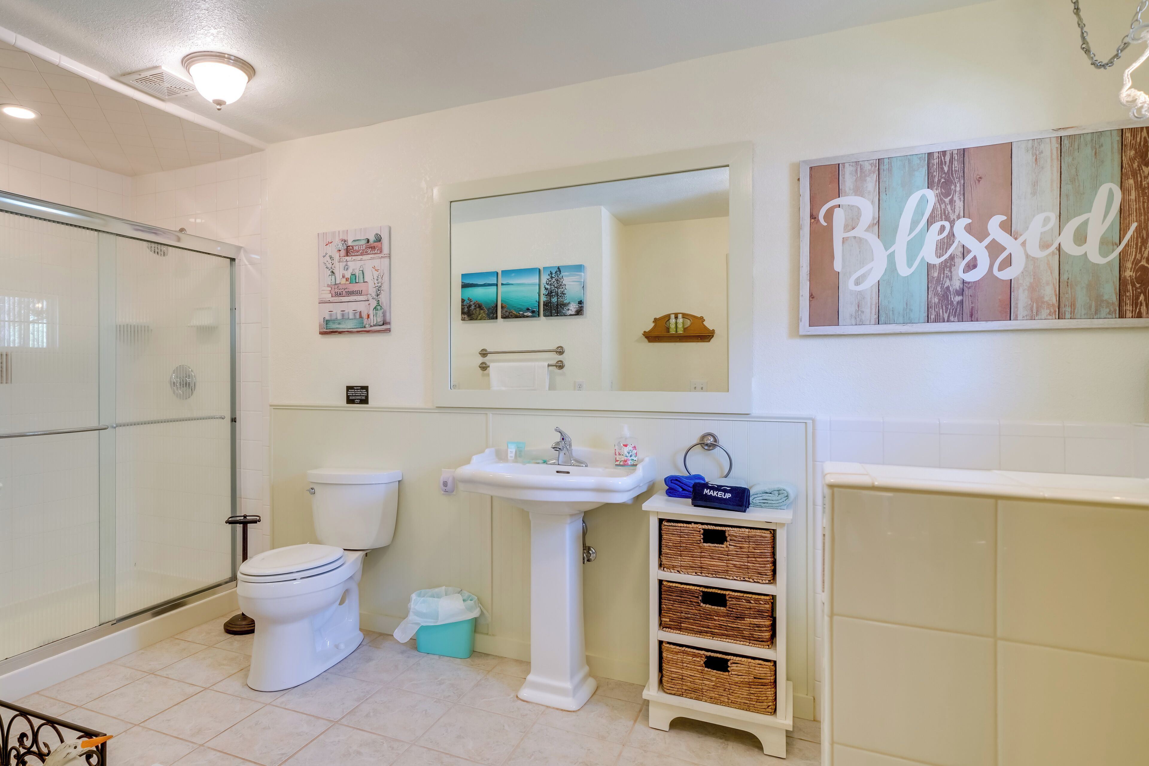 Une salle de bain avec une douche à parois de verre, un lavabo sur pied blanc, des toilettes et des paniers en osier posés sur un meuble-vasque.