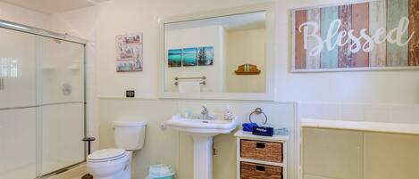 Une salle de bain avec une douche à parois de verre, un lavabo sur pied blanc, des toilettes et des paniers en osier posés sur un meuble-vasque.
