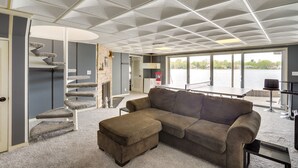 A living room with a large window, a sofa, a coffee table, and a spiral staircase. - Riverfront Elkhart Rental: Deck & Ping Pong Table! (Elkhart)