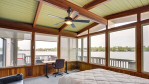 A room with a bed, a desk, and a chair, large windows with a view of a body of water, and a ceiling fan. - Riverfront Elkhart Rental: Deck & Ping Pong Table! (Elkhart)