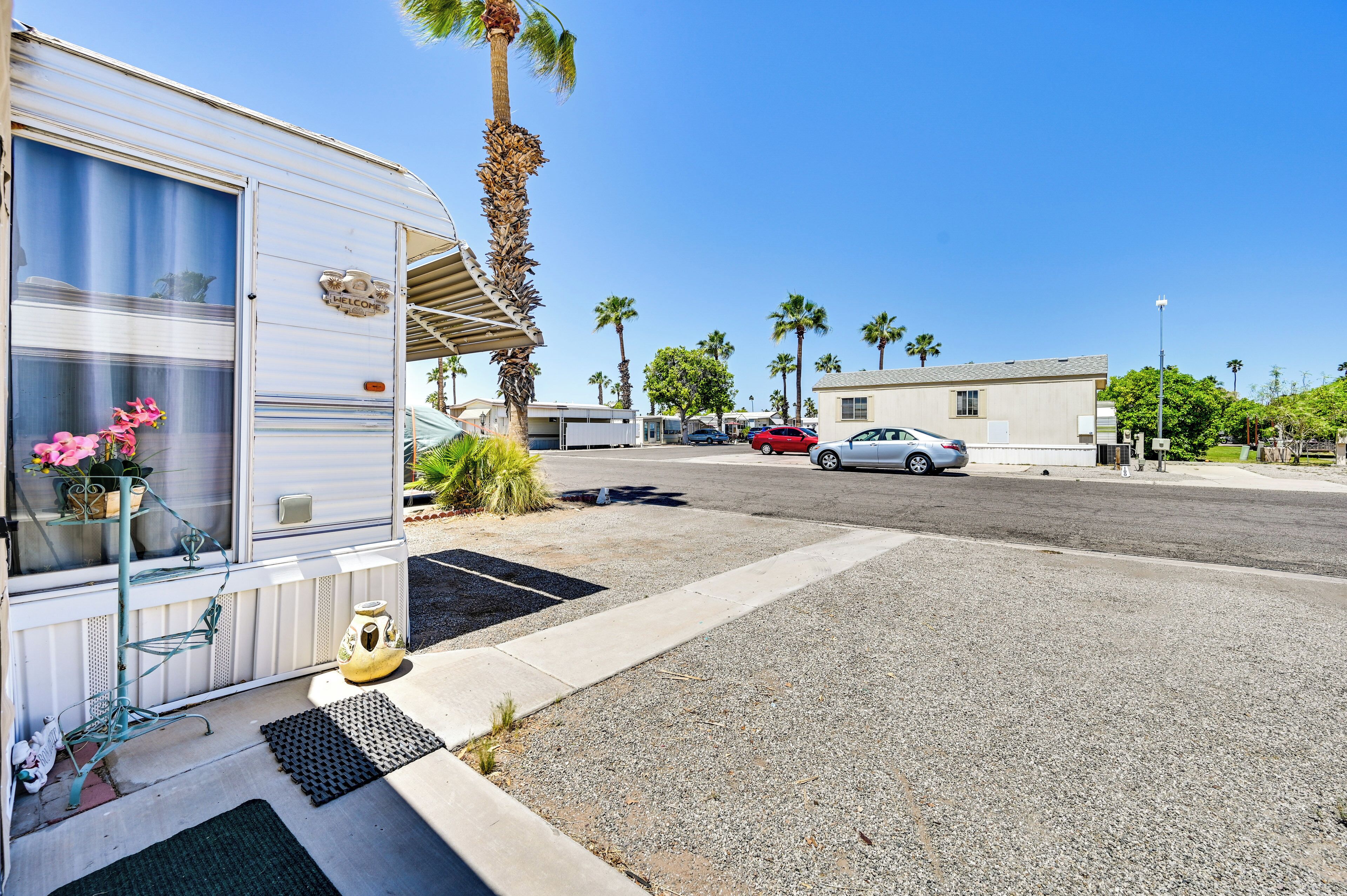 A mobile home with a covered porch, a welcome sign, and a small garden with pink flowers.