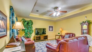 A living room with a brown leather sofa, a blue patterned rug, a ceiling fan, a television, and a clock on the wall. - 2 Mi to Uca: Group Getaway w/ Private Yard! (Conway)