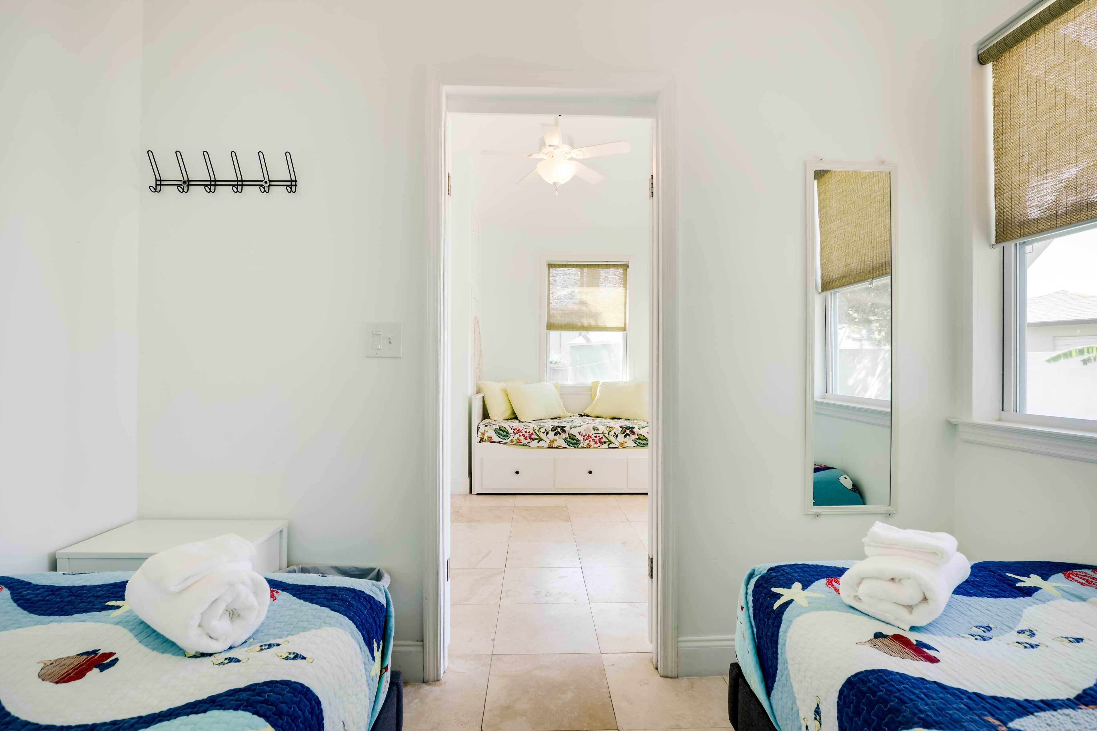 A hotel room with two beds, a ceiling fan, a window with a blind, and a hallway leading to another room.