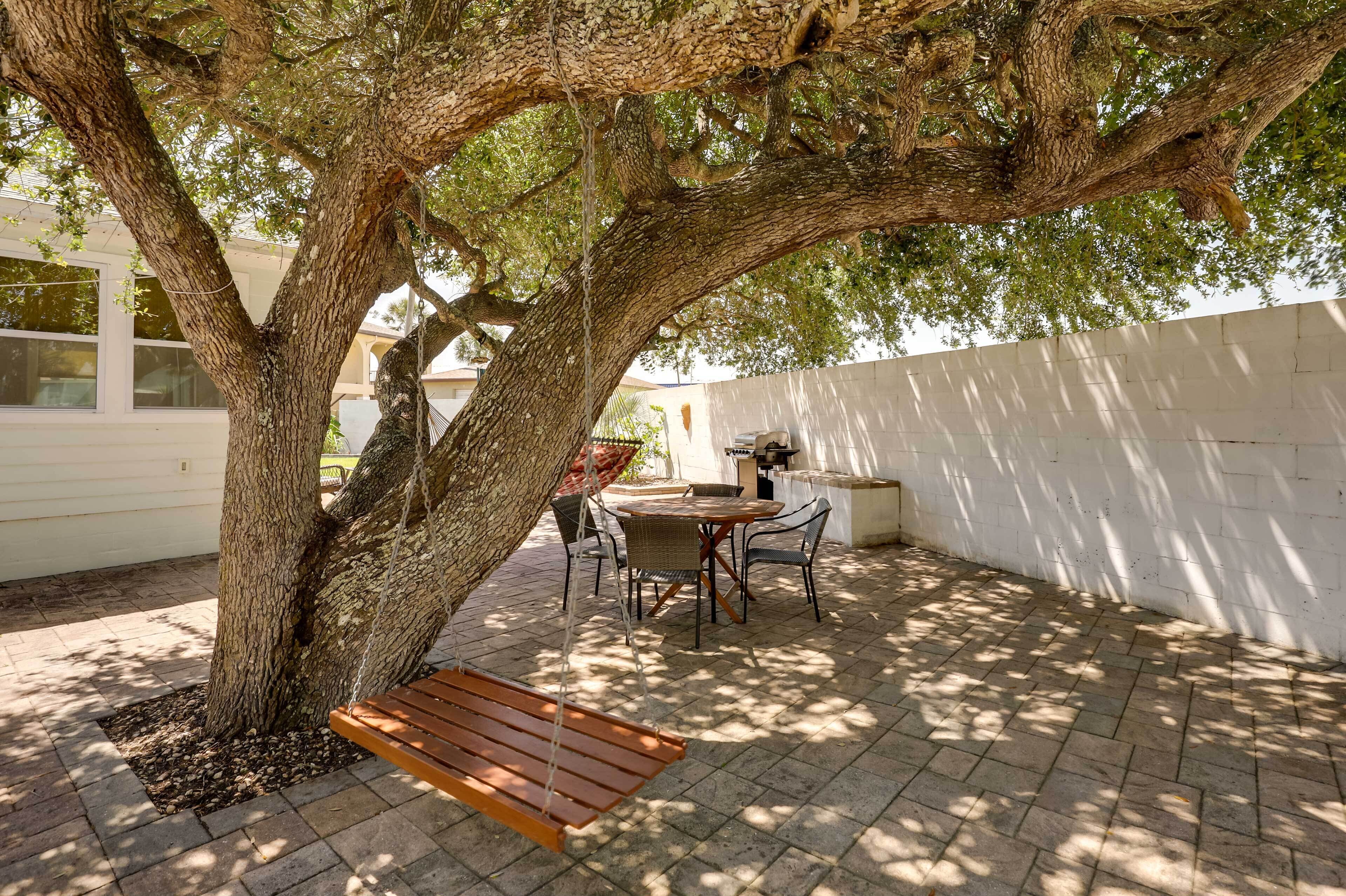 Un espace extérieur aménagé avec un grand arbre, une balançoire et une table entourée de chaises.