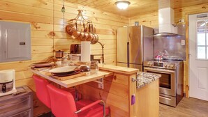 Une cuisine rustique dans une cabane en bois, avec une table en bois, une chaise rouge, des appareils électroménagers en acier inoxydable et un porte-casseroles suspendu.
