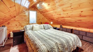 Une chambre confortable située sous les toits, avec un plafond en pente, un grand lit recouvert d’une literie à motifs, une table de chevet, une commode et une fenêtre laissant entrer la lumière naturelle.