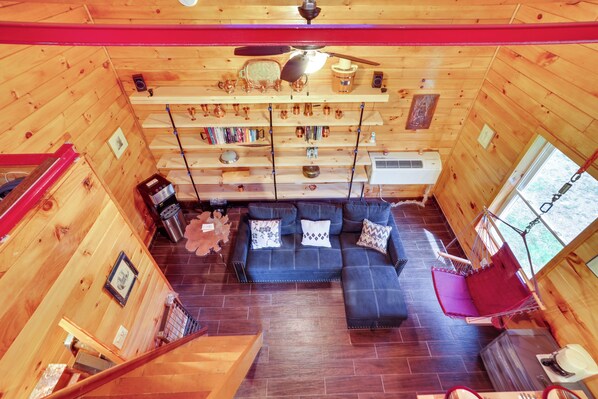 Un interior de cabaña acogedor, con piso de madera, un sofá negro con cojines estampados, un ventilador de techo y una ventana con vistas a los árboles.