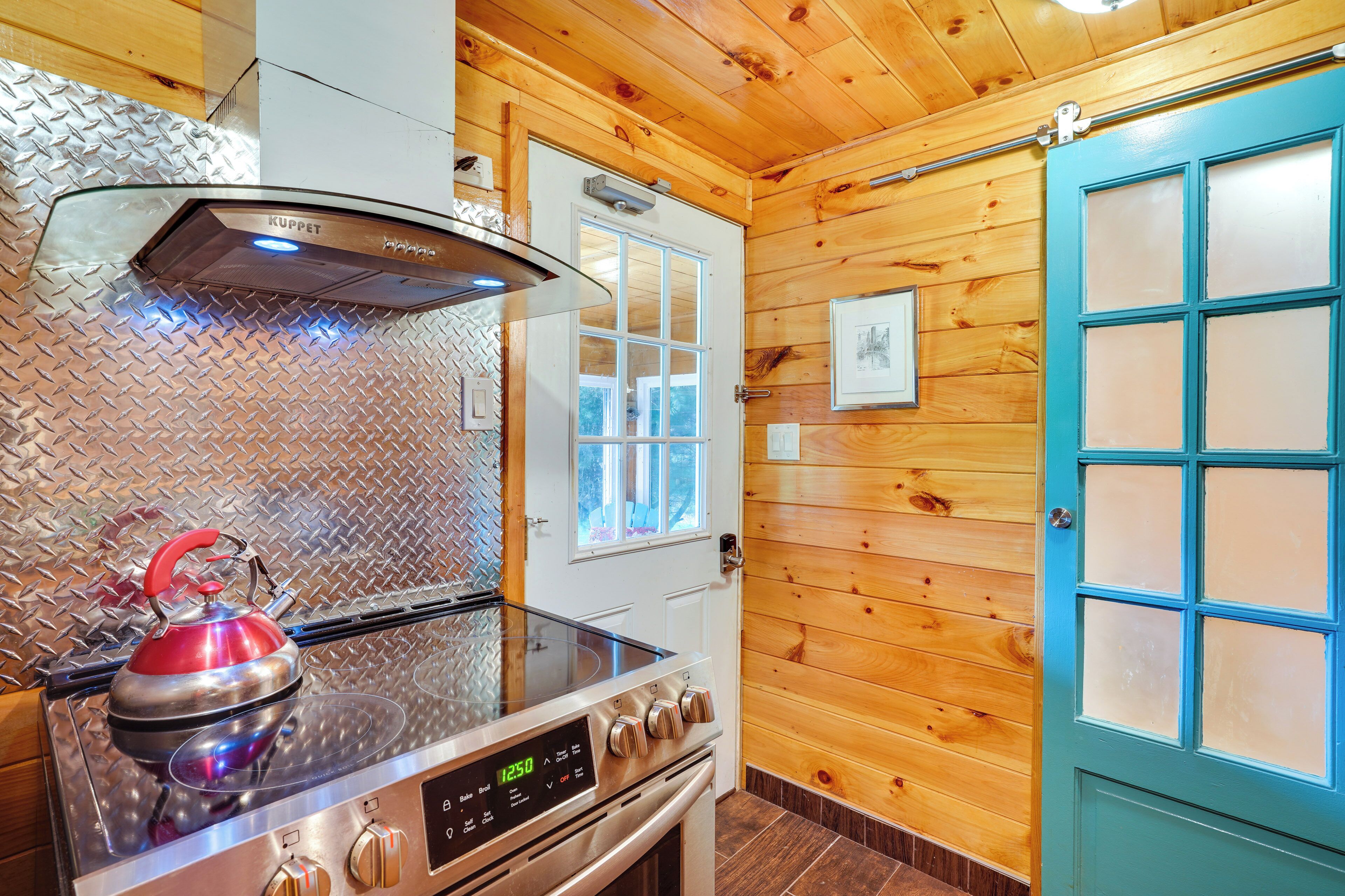A kitchen with a stainless steel stove, a red kettle, and wooden walls.