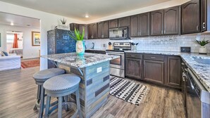 A modern kitchen with dark wood cabinets, a granite countertop, and stainless steel appliances. There are two bar stools and a potted plant on the island. - Boho Mountain Home Escape, Walk to Marina! (Mountain Home)
