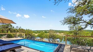 A swimming pool with lounge chairs and a wooden deck, surrounded by trees and a view of a lake and hills. - Updated Texas Tiny Home Rental on Lake Travis (Spicewood)