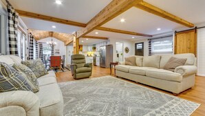 A spacious living room with a high ceiling and exposed wooden beams, a large sofa, a matching armchair, and a patterned rug. - Lakefront Arkansas Abode - Deck, Grill & Fire Pit (Scranton)