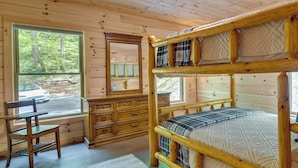 Ein Schlafzimmer einer Holzhütte mit Etagenbett, Kommode, Stuhl und Fenster mit Blick auf eine Straße und Bäume.