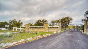 Une entrée fermée par un portail, avec une route goudronnée menant à un bâtiment, lequel est entouré de murs en pierre et d’une clôture blanche.