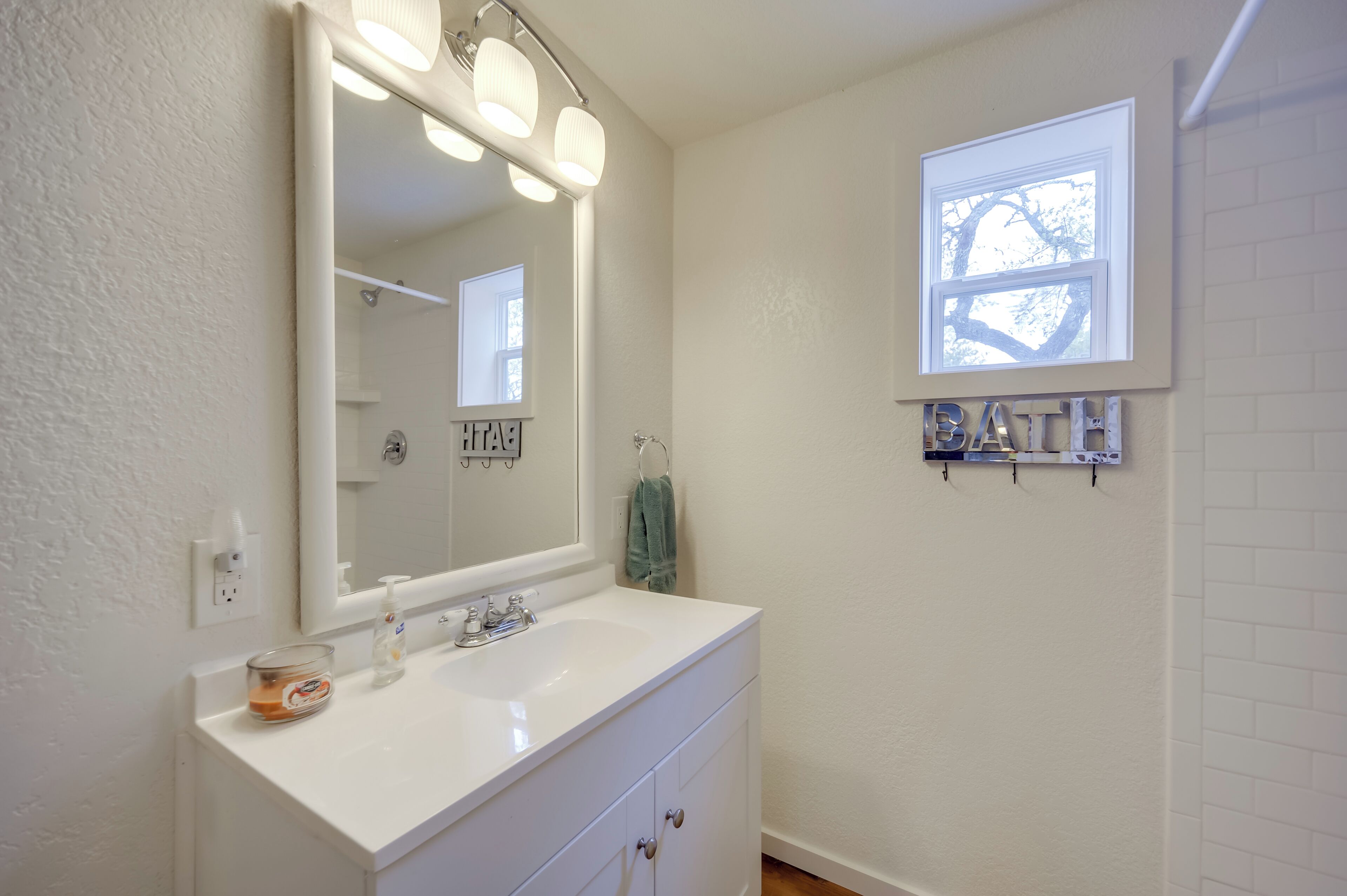 A bathroom with a white vanity, a large mirror with lights, a window, and a towel rack.
