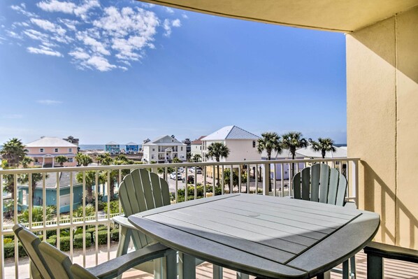 A balcony with a table and chairs, overlooking a coastal area with buildings and palm trees.