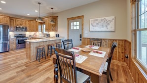 A spacious kitchen with wooden cabinets, a central island with stools, a dining table set for four, and a large window providing natural light. - Smoky Mountain Cabin Rental: Game Room, Fire Pit! (Andrews)