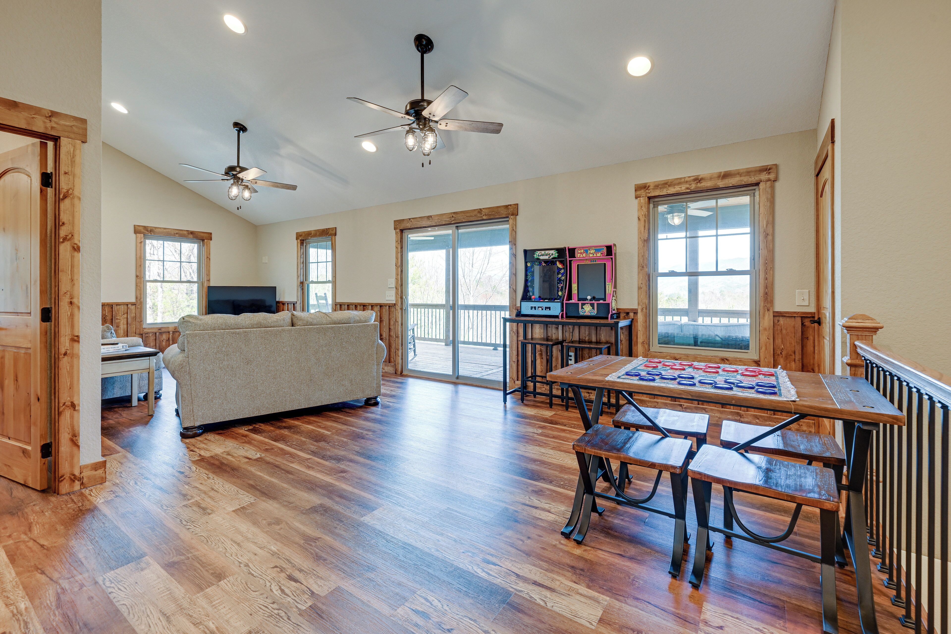 A spacious room with hardwood flooring, a ceiling fan, a sofa, a dining table with benches, and a large window with a view.