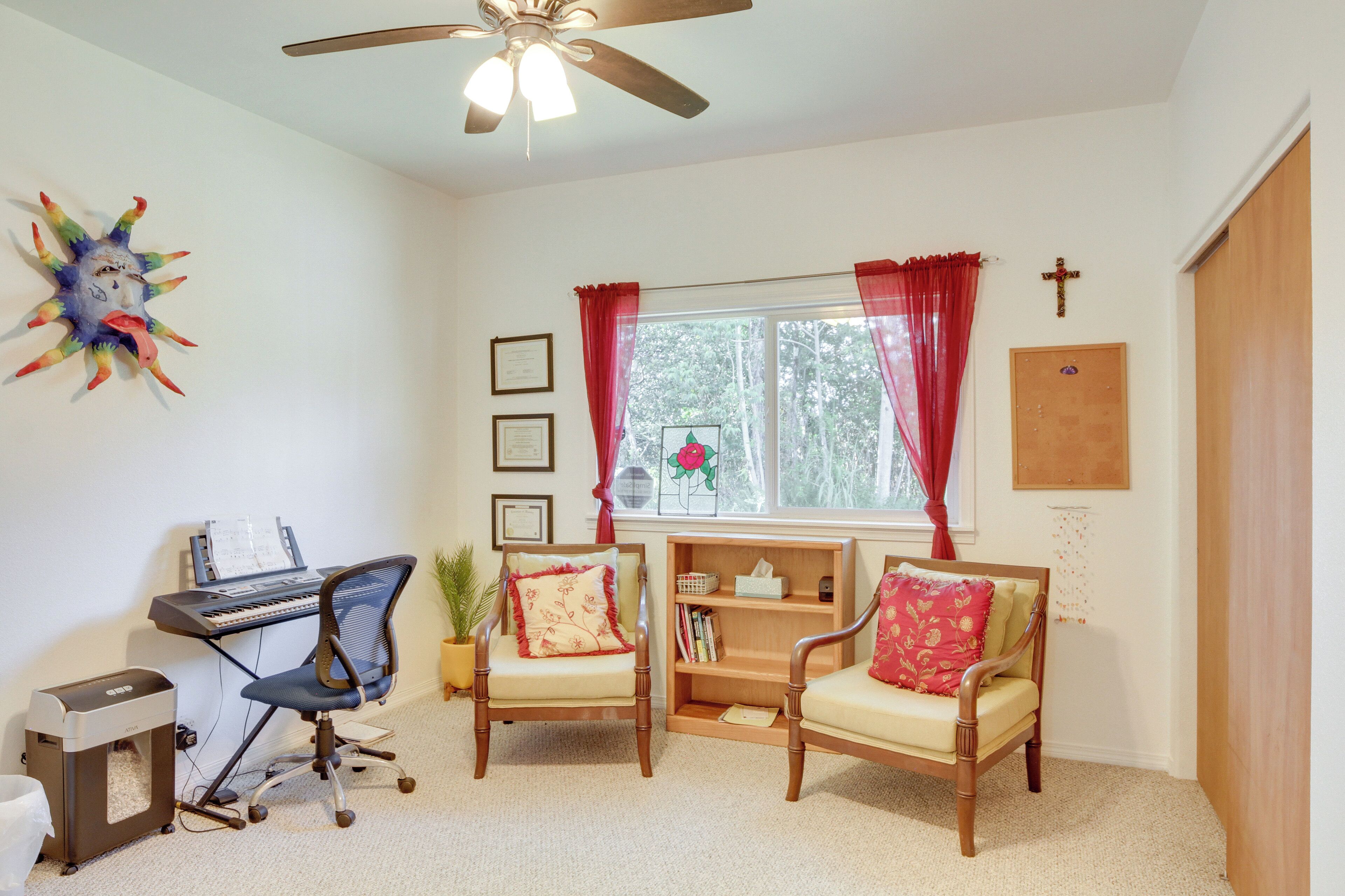 A room with a desk, two chairs, a ceiling fan, a window with curtains, a bookshelf, and a cross on the wall.